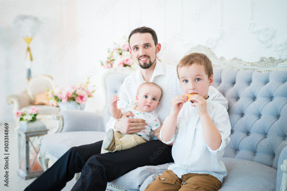 Positive young father is sitting on the blue couch with his little sons in a beautiful designer room. The concept of procreation and family. Father and son concept Positive young father is sitting on the blue couch with his little sons in a beautiful designer room. The concept of procreation and family. Father and son concept