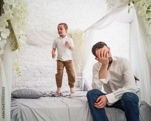 Tired young father is sitting on the edge of a large and beautiful bed where his little fidgety son is jumping. Concept of hyperactive children and tired parents photo