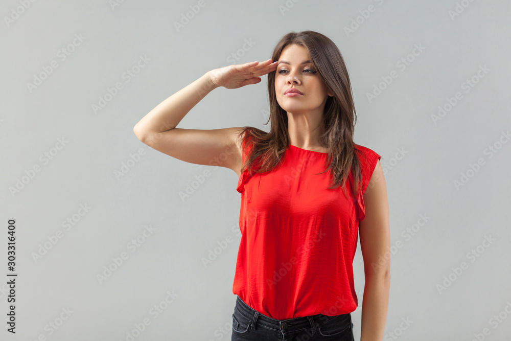 Yes sir! Portrait of serious beautiful brunette young woman in red shirt standing in salute mode and looking attentive and ready to perform the task . indoor, studio shot, isolated on gray background. Yes sir! Portrait of serious beautiful brunette young woman in red shirt standing in salute mode and looking attentive and ready to perform the task . indoor, studio shot, isolated on gray background.