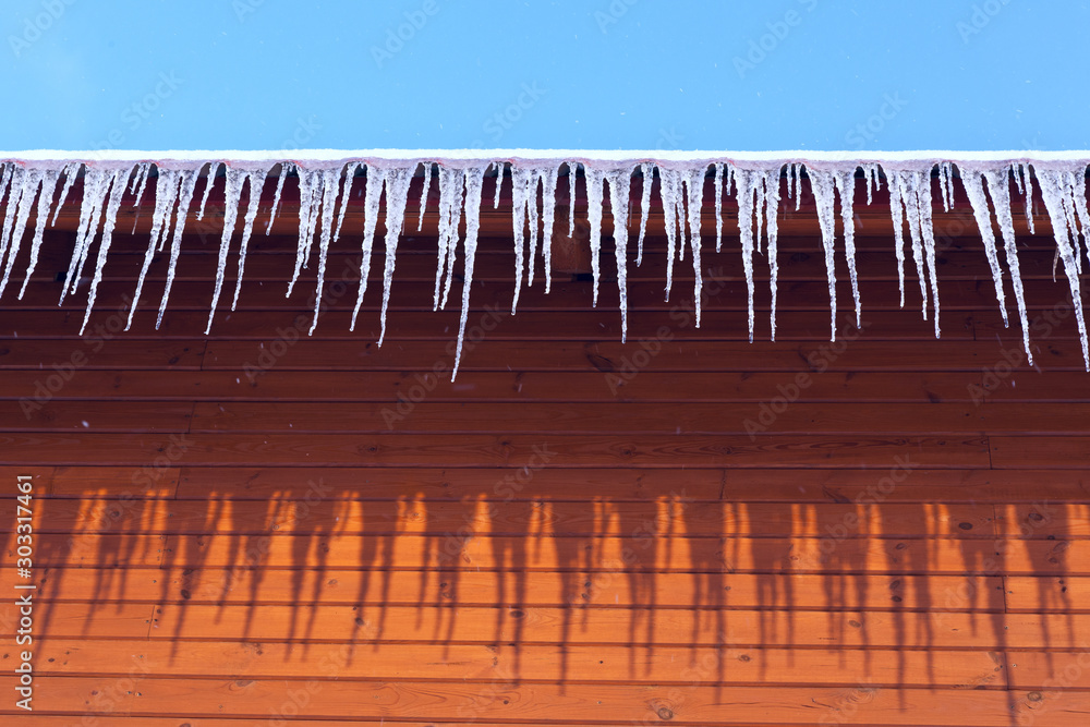 Icicles on the roof of the house against the background of the blue sky and the orange wall Icicles on the roof of the house against the background of the blue sky and the orange wall