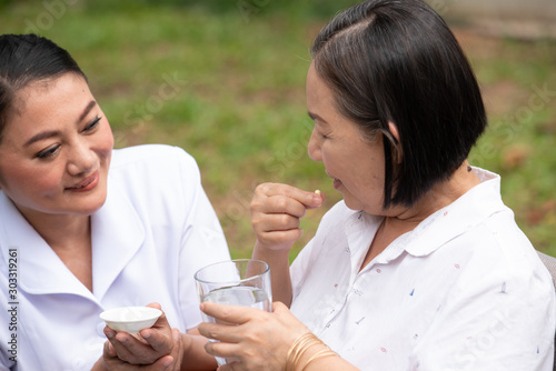 Asian nurse helping sick elderly woman to drink water with medical pill.