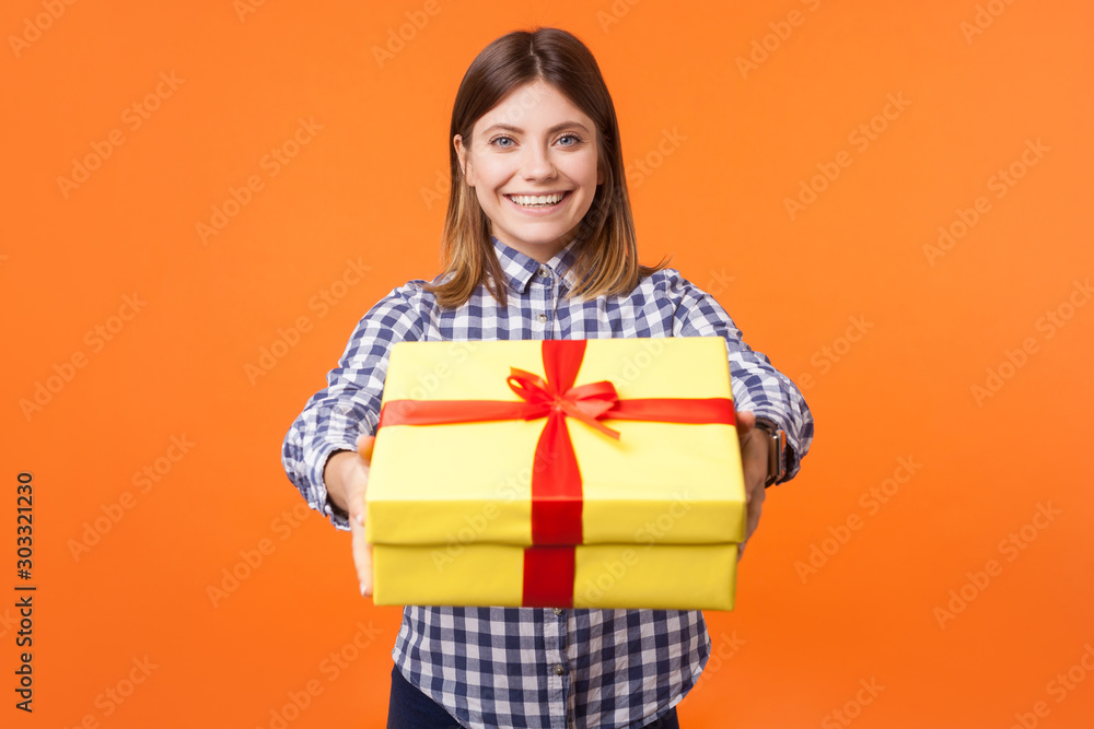 Portrait of generous friendly young woman with brown hair in casual checkered shirt looking at camera with toothy smile and giving gift box to camera. indoor studio shot isolated on orange background Portrait of generous friendly young woman with brown hair in casual checkered shirt looking at camera with toothy smile and giving gift box to camera. indoor studio shot isolated on orange background