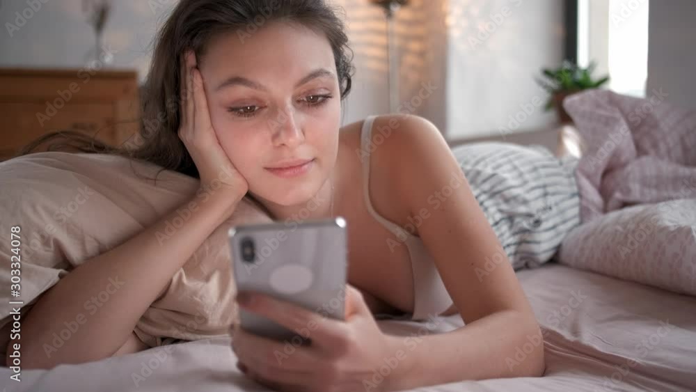 Relaxed woman at home reading a text message in her bright bedroom. Woman in bed checking social apps with smartphone Relaxed woman at home reading a text message in her bright bedroom. Woman in bed checking social apps with smartphone