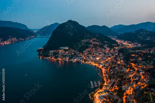 High angle aerial drone night shot of city street lights by lake monte salvatore in Lugano, Switzerland
