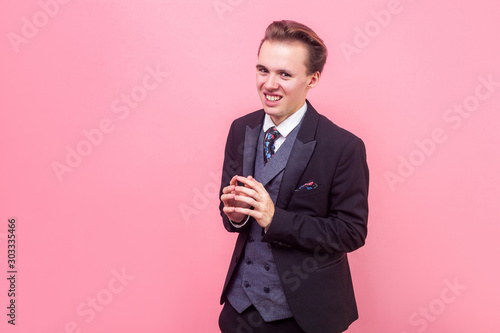 Portrait of devious sneaky sly businessman in suit and with stylish hairdo looking at camera with tricky cunning smile, thinking evil idea, meanness. indoor studio shot isolated on pink background photo