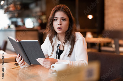 young beautiful woman in white jacket checking her planner working outside office drinking hot coffee in the trendy cafe looking confused multitasking modern businesswoman
