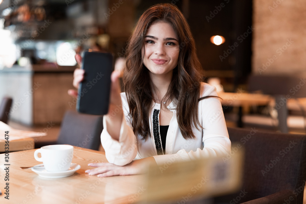 young smiling woman showing her smartphone during lunch break drinking hot coffee in trendy cafe looking happy modern businesswoman