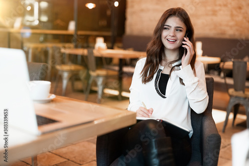young beautiful woman in white stylish jacket with smartphone in her hands looking pleased while speaking with friend during coffee break in cafe modern businesswoman
