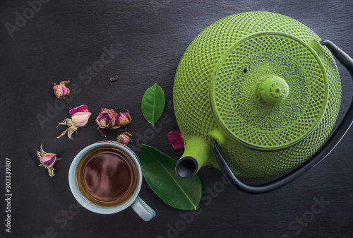 Tea time concept. Green iron teapot, cup of tea, rose buds and tea leafs ont he slate background. Copy space, Flat lay photo