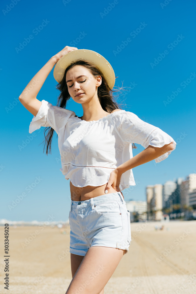 girl in a country style hat and blouse with an expression of determined despair, closing her eyes holds her hat on the background of the beach and buildings girl in a country style hat and blouse with an expression of determined despair, closing her eyes holds her hat on the background of the beach and buildings