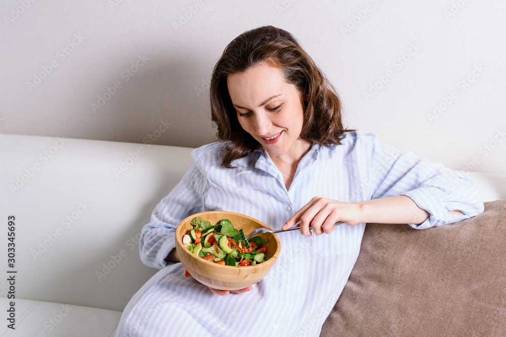 pregnant smiling woman sitting on sofa eating healthy vegetable salad in wooden bowl pregnant smiling woman sitting on sofa eating healthy vegetable salad in wooden bowl