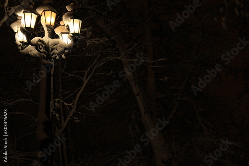 Night illumination of winter park in the central part of the town. Old-fashioned city lantern, covered with snow on the black background. Merry Christmas and Happy New Year