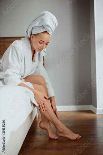 Beautiful happy woman sitting on bed in luxury hotel, having satisfied expression, looking down, examining skin on long slender legs after procedure of epilation, body care and treatment concept