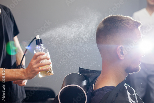 young Kazakh barber works in a barbershop, a young guy makes a short haircut at a hairdresser, wet his head, water splashes on a gray wall background