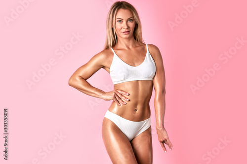 Portrait of blonde model in beautiful bra and penties standing against pink background in white romantic lingerie, keeping hand on hips, looking straight with positive expression, indoor shot