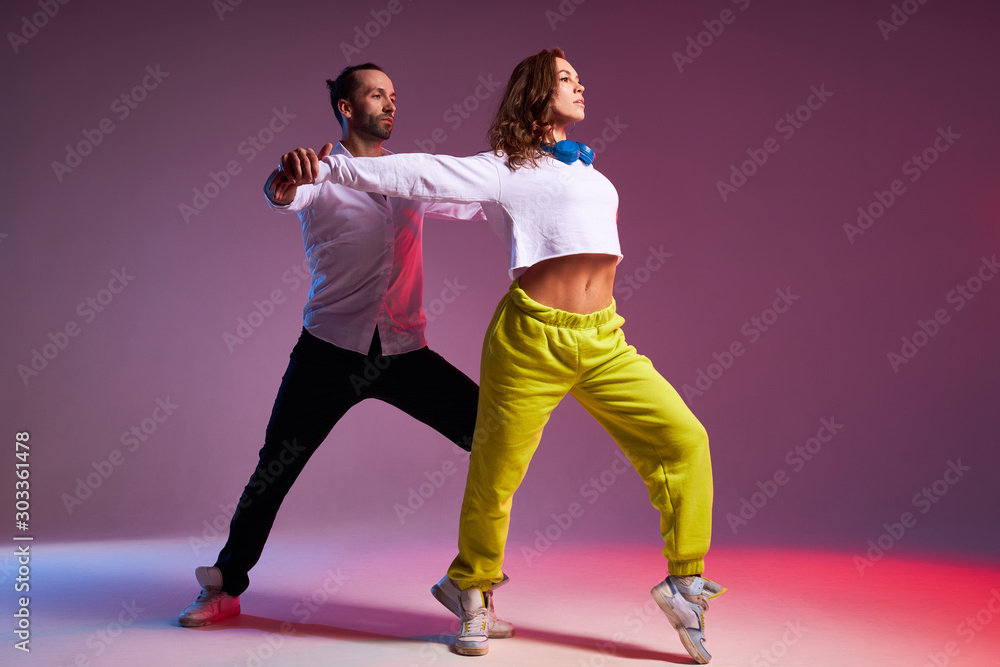 Charming young woman with long wavy hair, dressed in casual outfit dancing with active beard man in studio full of light, standing on tiptoe, looking away, dancers of soul disco concept