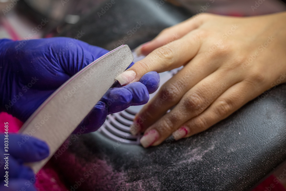 Manicurist with blue gloves rasping nails on women's hand in nail salon.