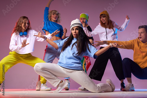 Teenage girl dancing hip-hop on modern dance studio, sitting on floor in white pants, spreading hands up, looking straight, training before concert