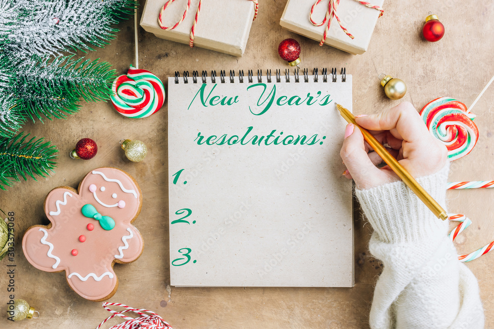 Obraz premium Notebook in the center of cute Christmas decoration: gift box, christmas balls, gingerbread man, fir tree branch, cande cane. Female hand writing in notebook. Flat lay. Top view