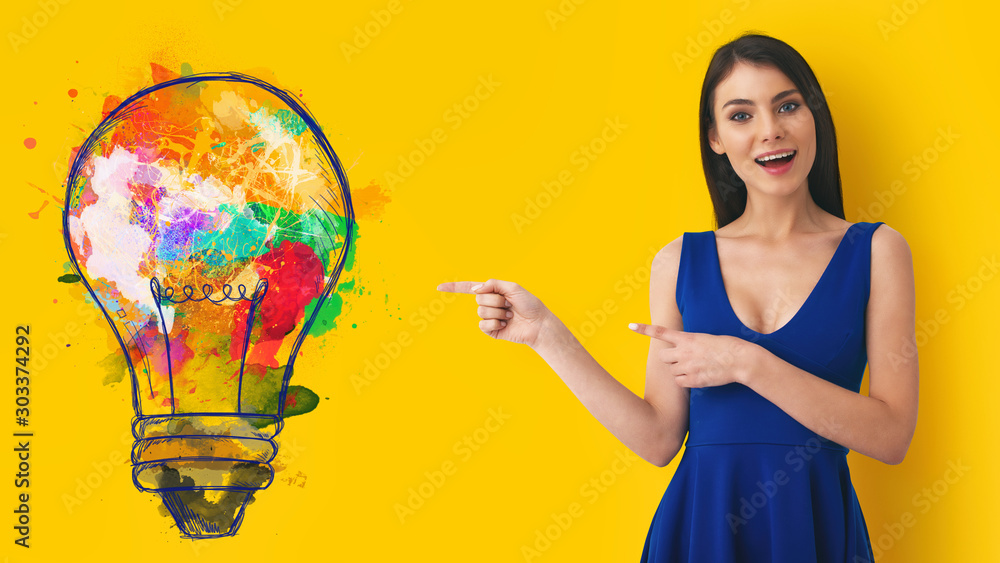 Young woman pointing at big stylized light bulb on yellow background. Concept of idea and creativity - obrazy, fototapety, plakaty 