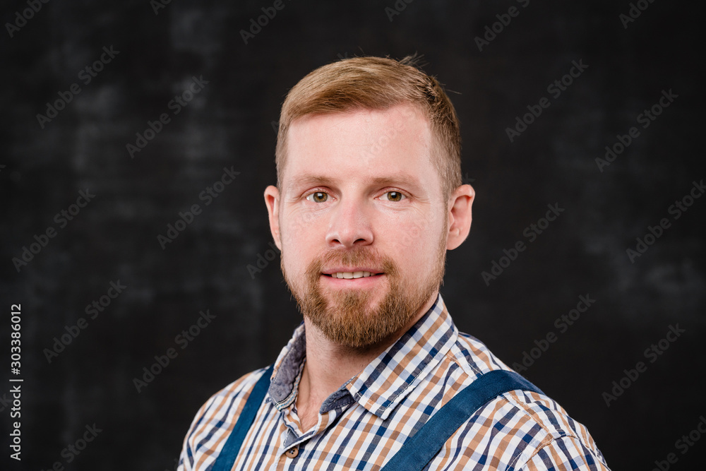 Young bearded technician in checkered shirt and blue overalls looking at you
