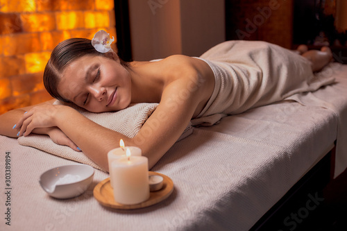 Beautiful young lady lying down on her belly in spa salon, getting massage on her back, relaxes woman with flower on head, candles near her.