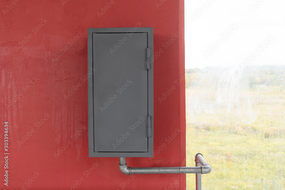 A gray iron box hangs on a red wall, next to a window. Fire box or gas box, a water or gas pipe is connected.