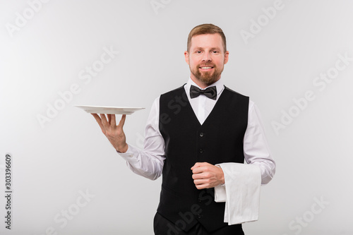 Young elegant waiter in black waistcoat and bowtie holding white towel and plate