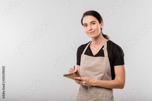 Foto Pretty young brunette waitress in apron making notes about order in notepad