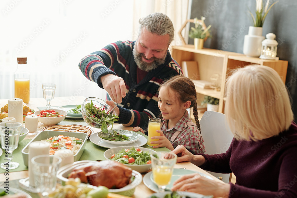 Happy grandpa giving salad to his cute granddaughter by festive table