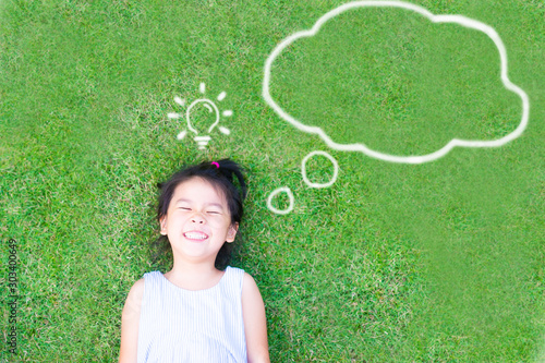 Smart asian girl lay down on green grass and thinking and discover good creative idea.World thinking day concept with girl kid having fun with dream and imagination cloud and light bulb idea. photo