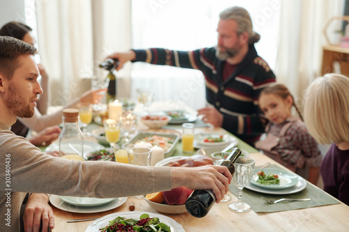 Young man pouring wine into glass of mom sitting in front of him during dinner