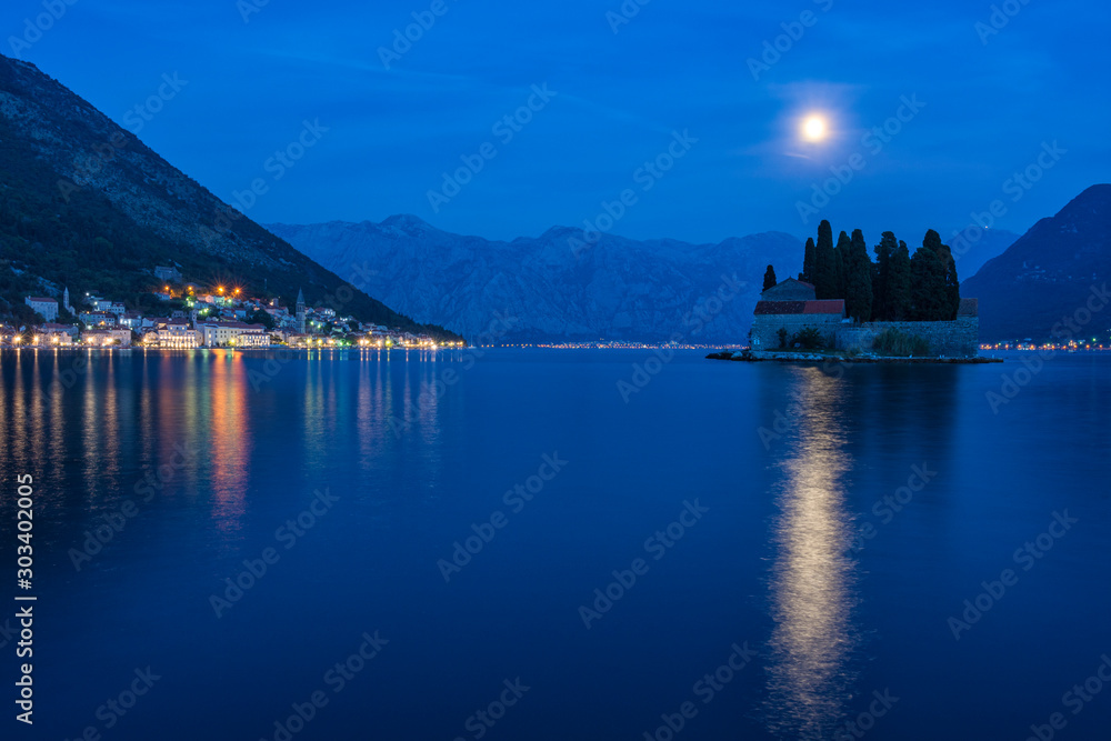 Lights of the ancient town of Perast (Montenegro) at night, reflecting on water. Island of Saint George litted up by the rising Moon. Bay of Kotor at blue hour. Cityscape at night, background.