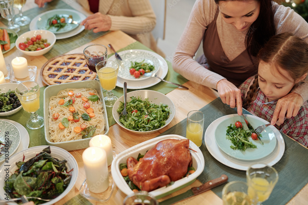 Young mother cutting tomato on plate of her little daughter by family dinner Young mother cutting tomato on plate of her little daughter by family dinner