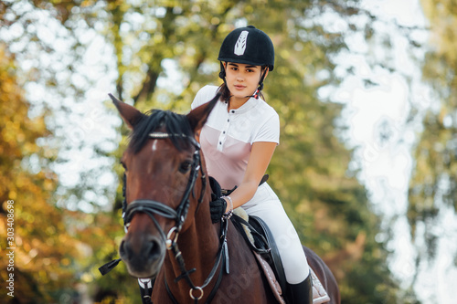 Portrait of young jockey girl riding a brown horse in autumn forest.