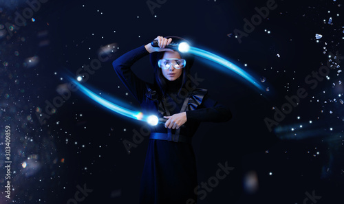 Beautiful woman in futuristic dress over dark space magic background. Gamer girl in glasses of virtual reality with controllers in hands. Augmented reality, game, hobby concept. VR. Blue neon light.