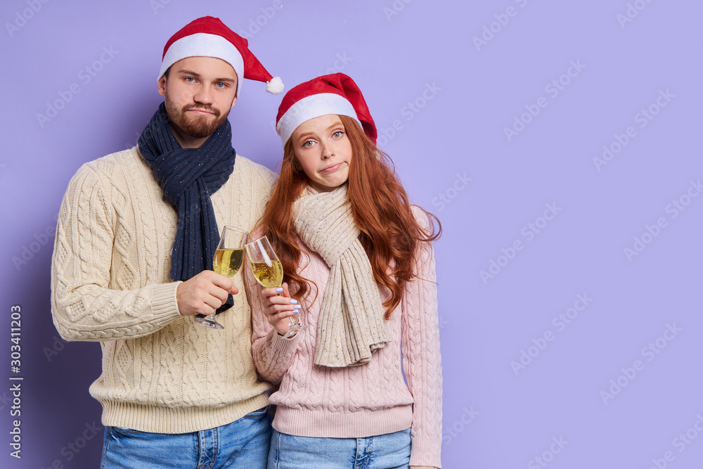 Tired upset looking couple standing together in Santa's hat, holding glasses of champagne, having bothering party, waiting for taxi to go home, isolated on violet background, indoor shot Tired upset looking couple standing together in Santa's hat, holding glasses of champagne, having bothering party, waiting for taxi to go home, isolated on violet background, indoor shot