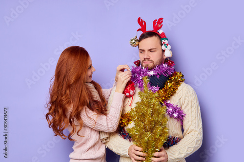 Dissatisfied, displeased man looking at chemung cheerful girl decorating him with different Christmas baubles, isolated family shut, posing over blue background, indoor shot photo
