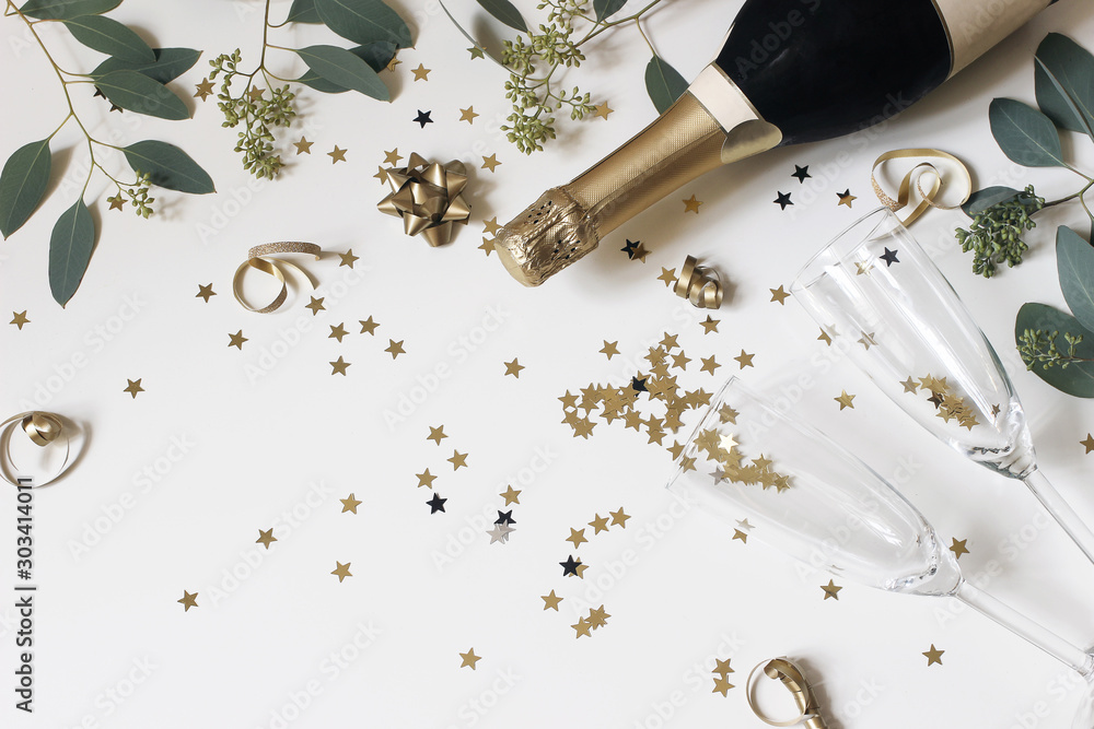 Happy New Year composition. Champagne glasses nad wine bottle with golden confetti stars and eucalyptus branches isolated on white table background. Celebration, party concept. Flat lay, top view. - obrazy, fototapety, plakaty 
