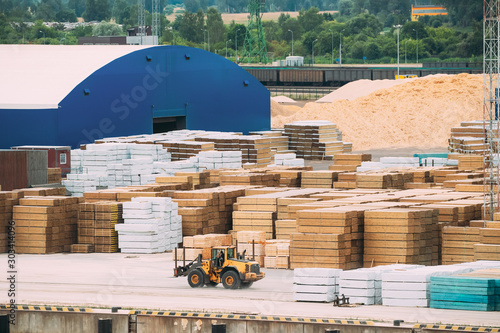 Industrial Forklift Drives Near Stacked Timber In Stock. Lift Truck, Jitney, Fork Truck, Fork Hoist, And Forklift Truck Working Near Industrial Lumber Material Storage Outdoors photo