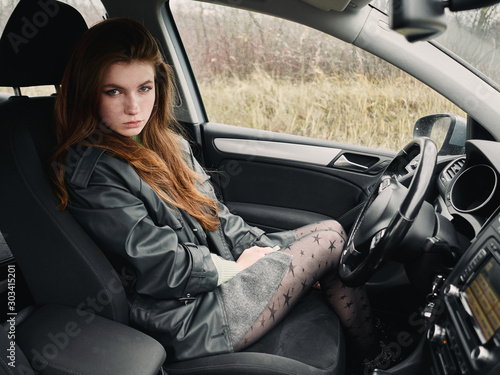 Close up portrait of sad young attractive red hair woman driver dressed black leather jacket grey skirt sitting in driver's seat cinematic vintage film filter colors © sergeyzapotylok