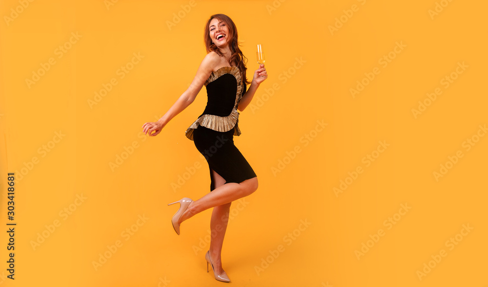 Full length image Young happy woman on yellow background drinking champagne, celebrating new year, wearing black dress, happy carnival disco party, sparkling confetti, holding glass, having fun. Full length image Young happy woman on yellow background drinking champagne, celebrating new year, wearing black dress, happy carnival disco party, sparkling confetti, holding glass, having fun.