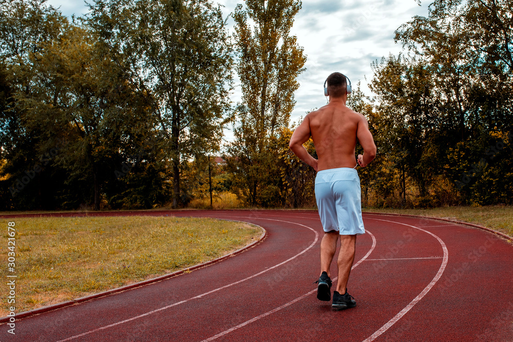 Back view of shirtless athlete man with headphones jogging on a track