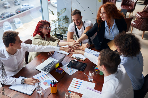 Positive group of co-workers joined hands together finishing up successful business meeting in conference office, papers, colorful pencils on table. Creative workers