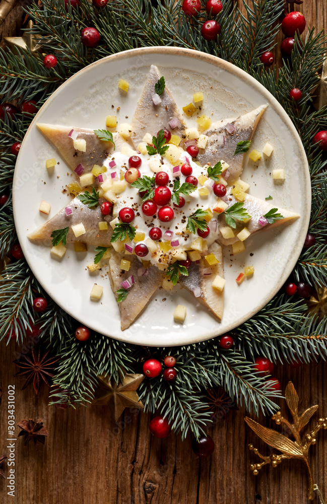 Christmas Herrings fillets with cream sauce with apple, pickled cucumbers, red onion and spices, garnished with cranberries on a ceramic plate on a festive decorated wooden table, top view