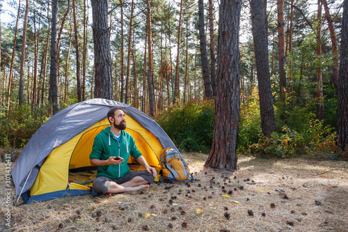 Handsome bearded young man listening to music in headphones while sitting in yellow tent in autumn forest. Hiking, travel, weekend trip concept. Healthy active lifestyle