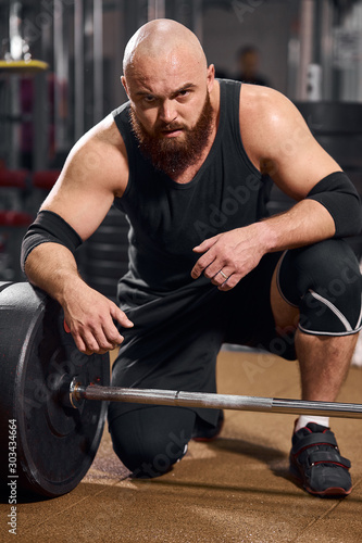 Side shot of hairless athlete with thick brunette beard, putting hand on heavy barbell, looking at camera with serious face, posing in gym hall, indoor shot