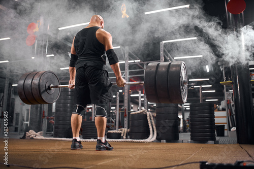 powerful man wearing black sporty uniform and professional sneackers, standing in front of heavy barbell with back to camera, looking down, bending head, preparing for deadlift, standing in smoke
