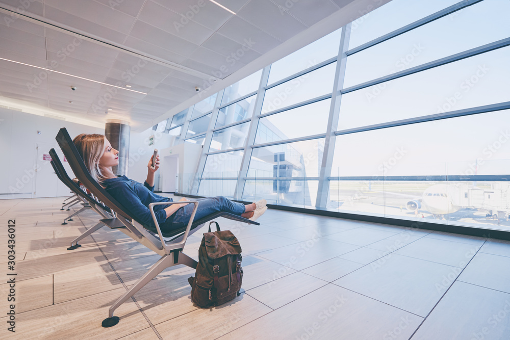 Obraz premium Travel and technology. Pretty young woman using smartphone while sitting at airport terminal waiting for boarding.
