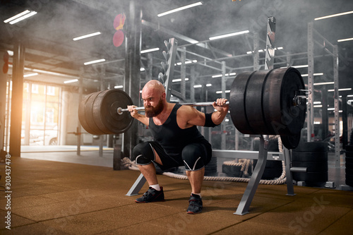 Sporty active man with strong arms and severe face, trying to stand with heavy barbell on hands, strong powerlifter training in gym, white smoke in the air, side indoor shot photo